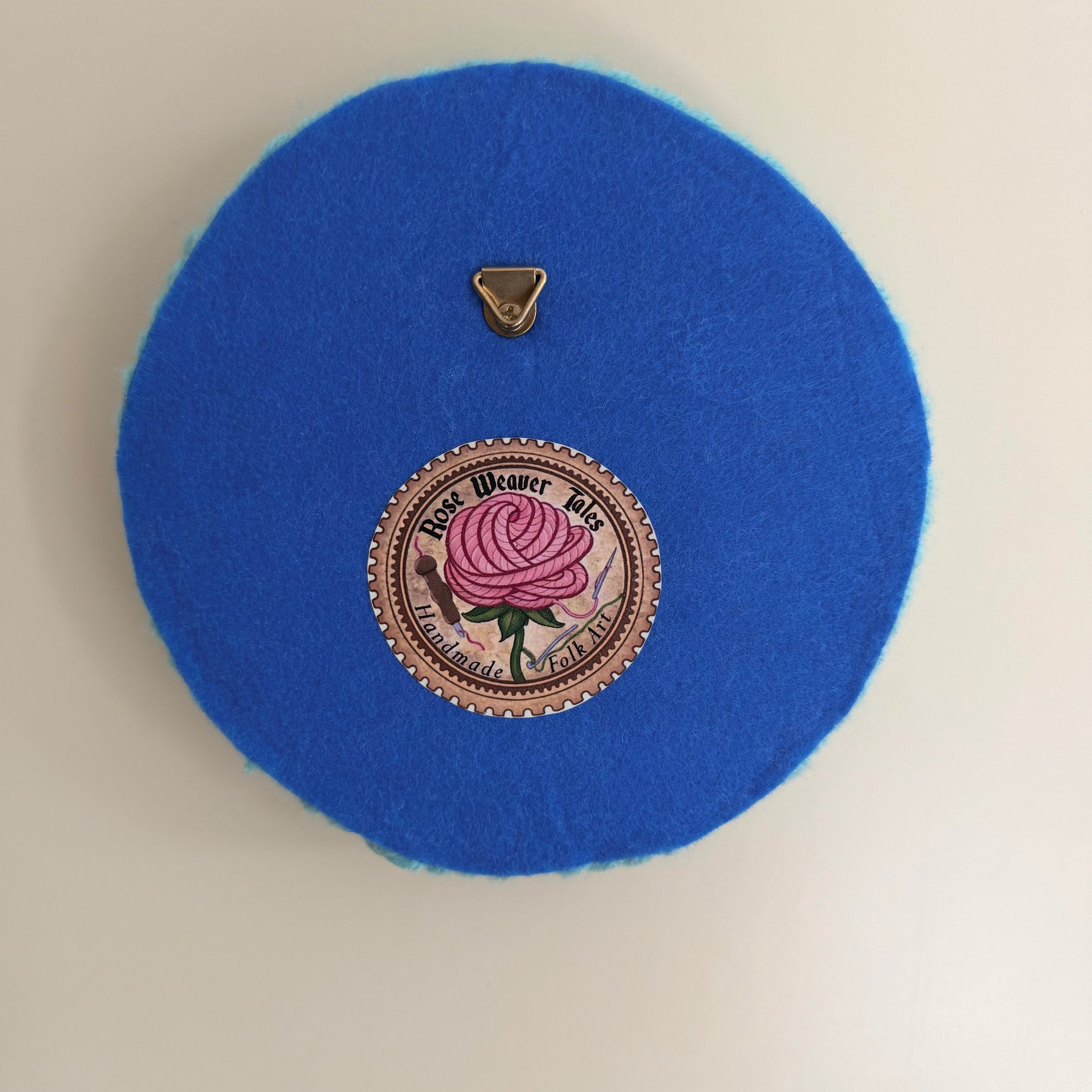 Back View of the handmade punch needle textile in round blue felt, showing a meat soft felt backing, a Rose Weaver tales brand label, and a segure metal hanging loop.