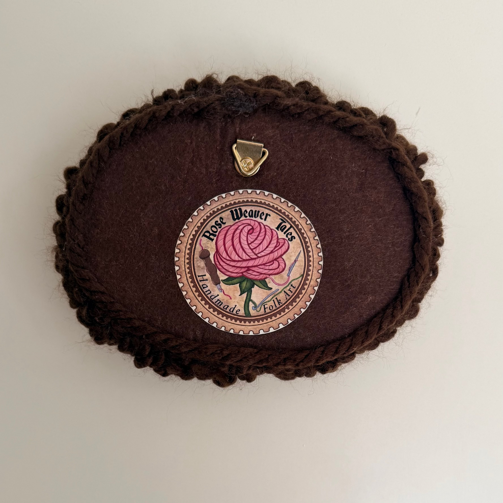 Back View of the handmade punch needle textile in brown felt oval, showing a meat soft felt backing, a Rose Weaver tales brand label, and a segure metal hanging loop.