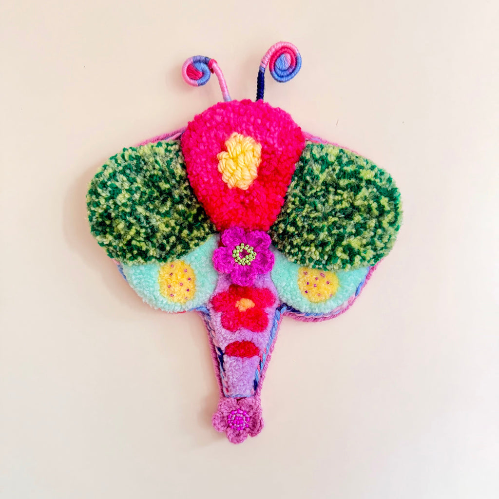 A colorful, handcrafted felt Dragonfly wall hanging on a plain off-white background. The dragonfly features green textured wings, a bright pink central body, yellow and purple flower details, and blue and pink antennae.