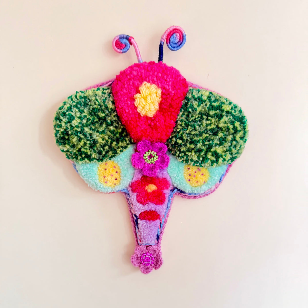 A colorful, handcrafted felt Dragonfly wall hanging on a plain off-white background. The dragonfly features green textured wings, a bright pink central body, yellow and purple flower details, and blue and pink antennae.