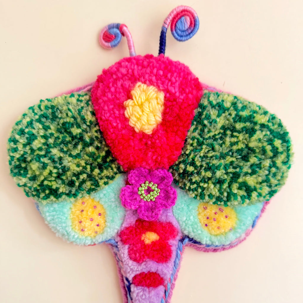 A colorful, handcrafted fuzzy Dragonfly wall hanging or ornament on a plain off-white background. The design features a bright pink fuzzy body with a yellow flower detail, green fuzzy wings, a light blue section, and purple antennae and tail.