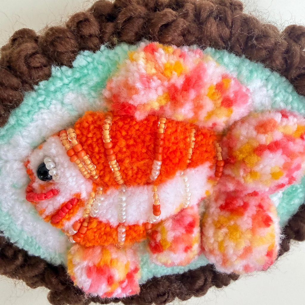 A close-up of a handcrafted felt fish ornament with orange, white, and pink marbled fur, decorated with small beads, set against a turquoise background with a brown curly border.