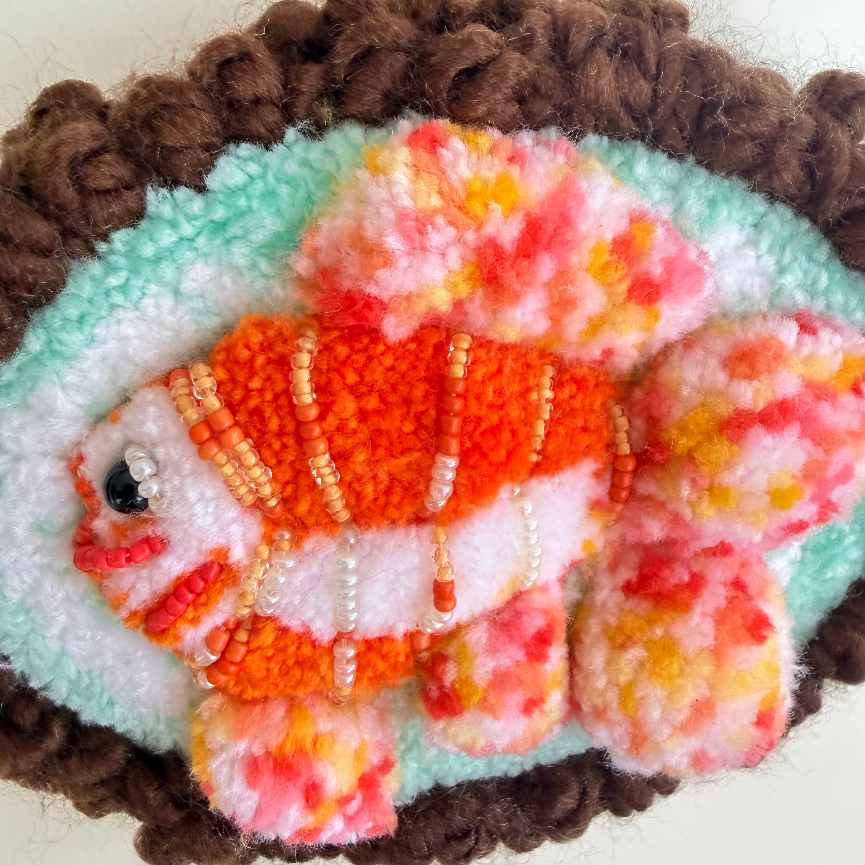 A close-up of a handcrafted felt fish ornament with orange, white, and pink marbled fur, decorated with small beads, set against a turquoise background with a brown curly border.