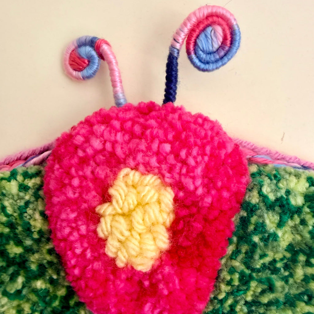 A close-up of a colorful, fuzzy handmade wall hanging or decorative piece featuring a bright pink flower with a yellow center, green textured wings, and pink, blue, and yellow antennae against a plain beige background.