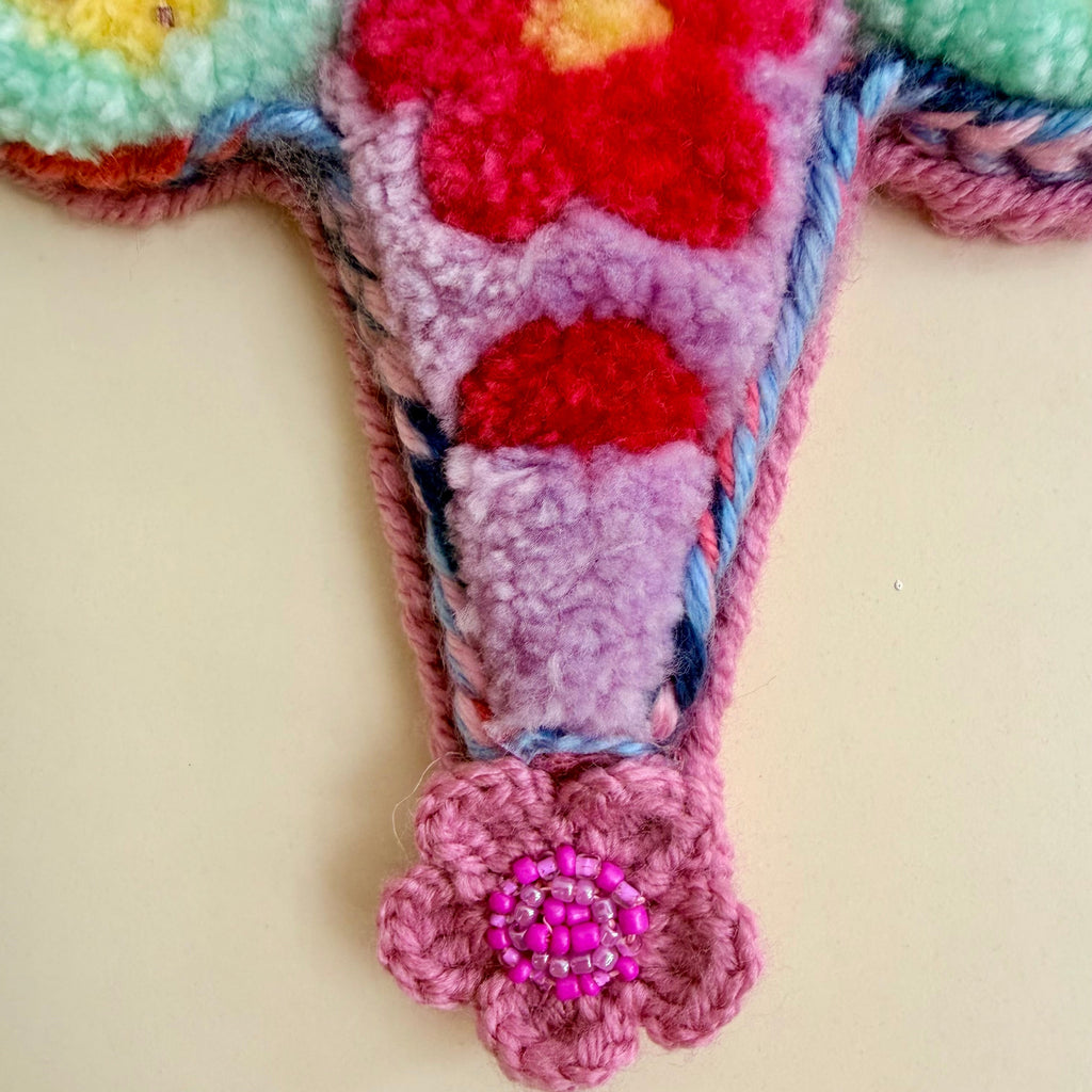 A close-up of a colorful, fuzzy crocheted and tufted decorative piece with a pink flower-shaped hanging element at the bottom, set against a plain beige background.