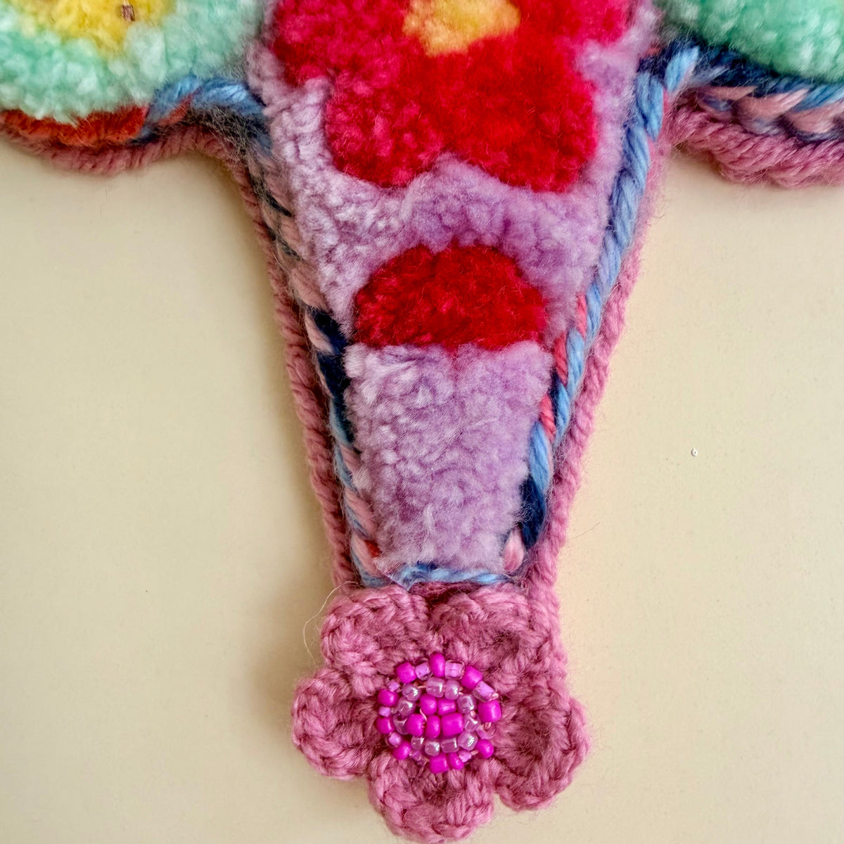 A close-up of a colorful, fuzzy crocheted and tufted decorative piece with a pink flower-shaped hanging element at the bottom, set against a plain beige background.