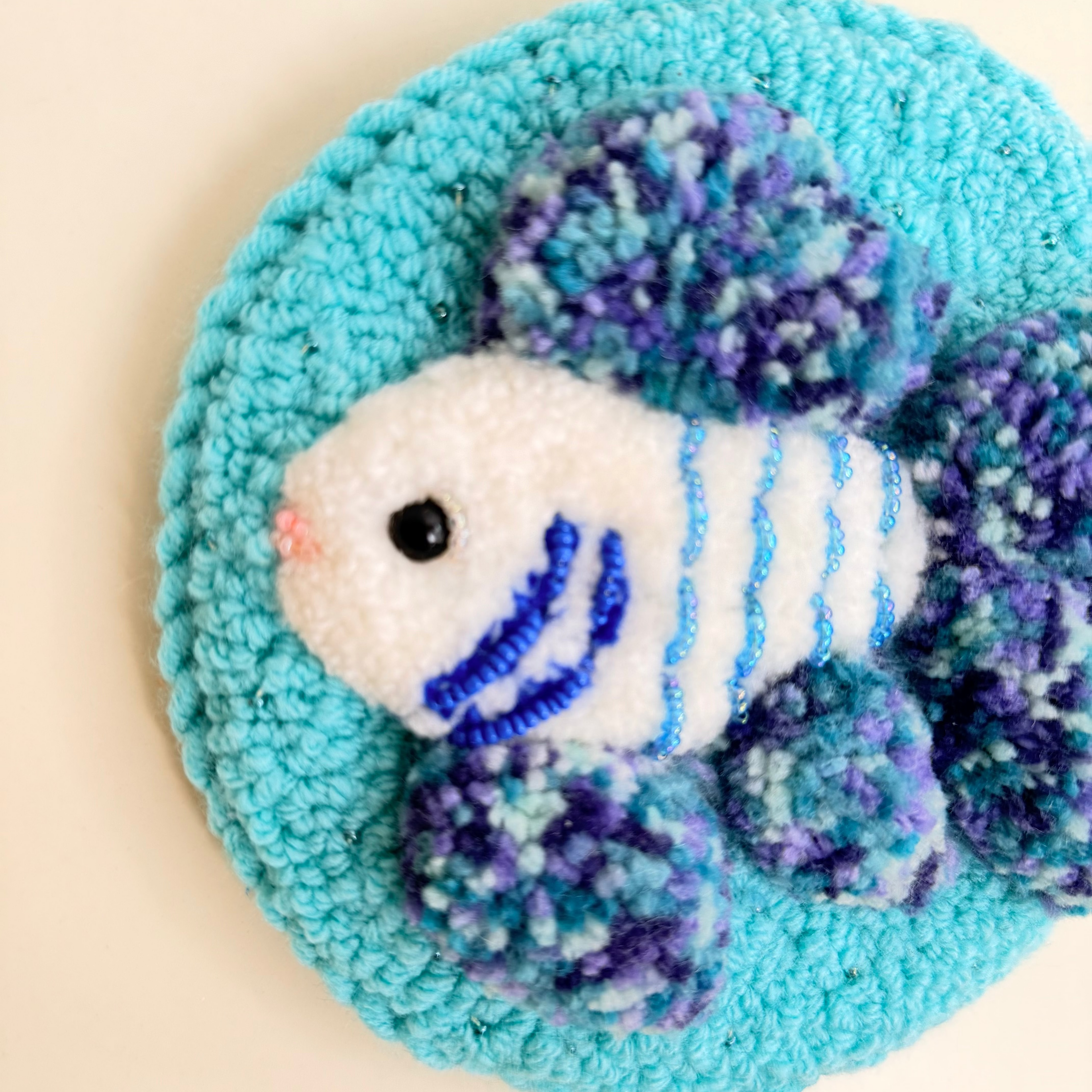 A handmade, fuzzy fish-shaped wall hook or decorative plaque on a circular turquoise textured base, set against a plain cream background.