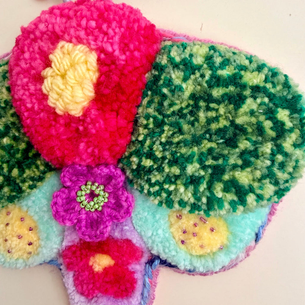 A colorful, handcrafted fuzzy dragonfly wall hanging made from textured yarn and faux fur, featuring bright pink, green, yellow, and purple colors on a plain light background.