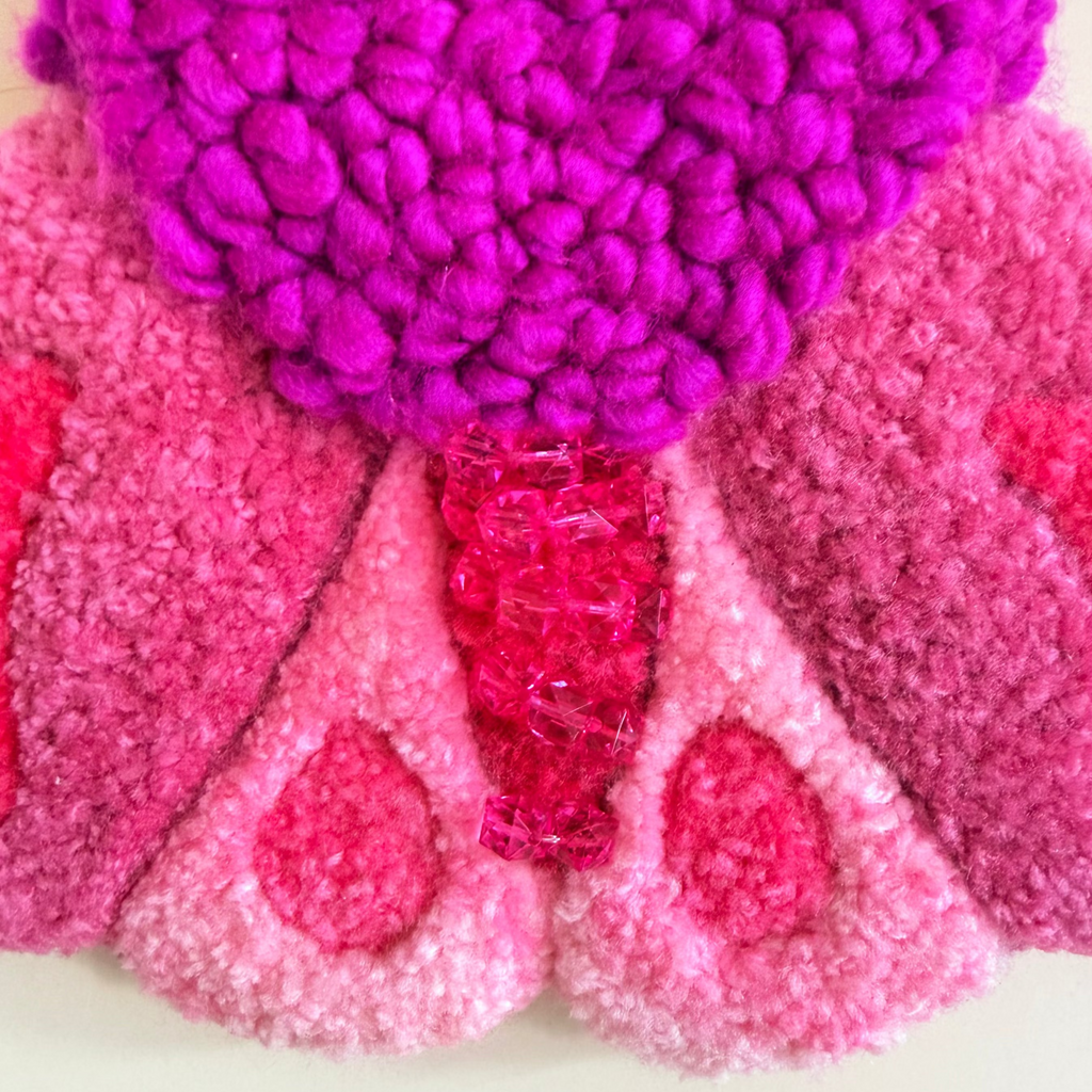 A close-up of a textured, wall hanging shaped like a stylized moth, made from punch needle, style yarn in shades of magenta, pink, and purple, mounted on a plain beige wall.