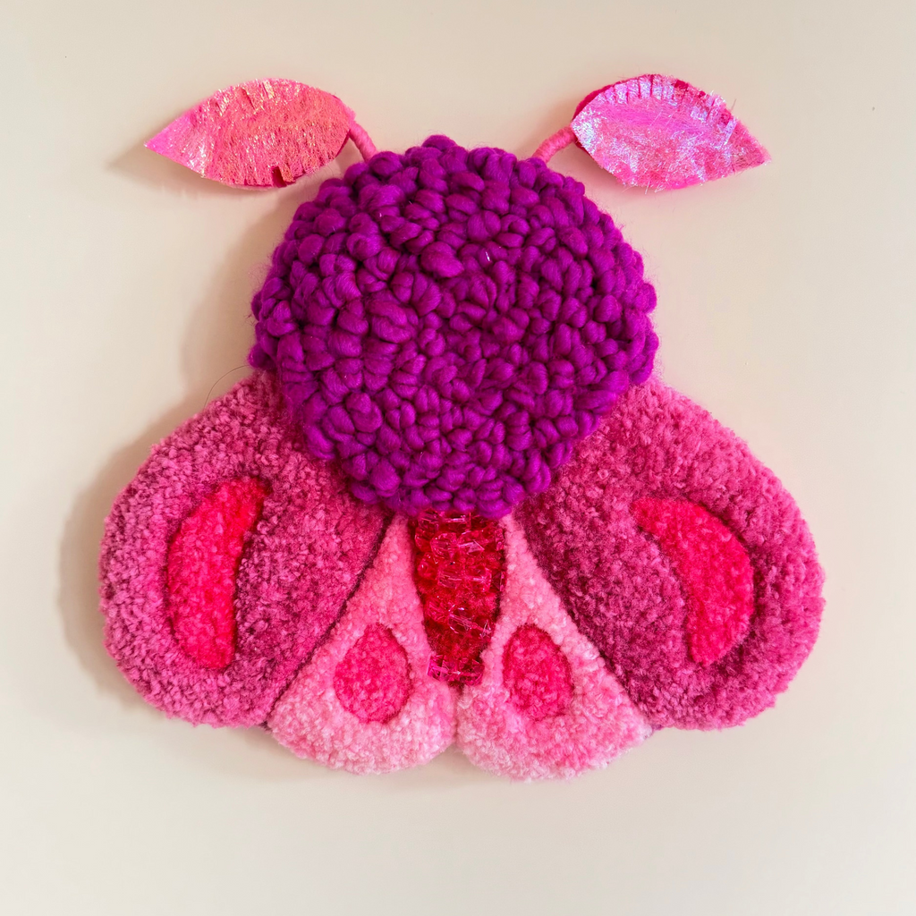 A colorful, textured wall hanging shaped like a stylized moth, made from punch needle, style yarn in shades of magenta, pink, and purple, mounted on a plain beige wall.
