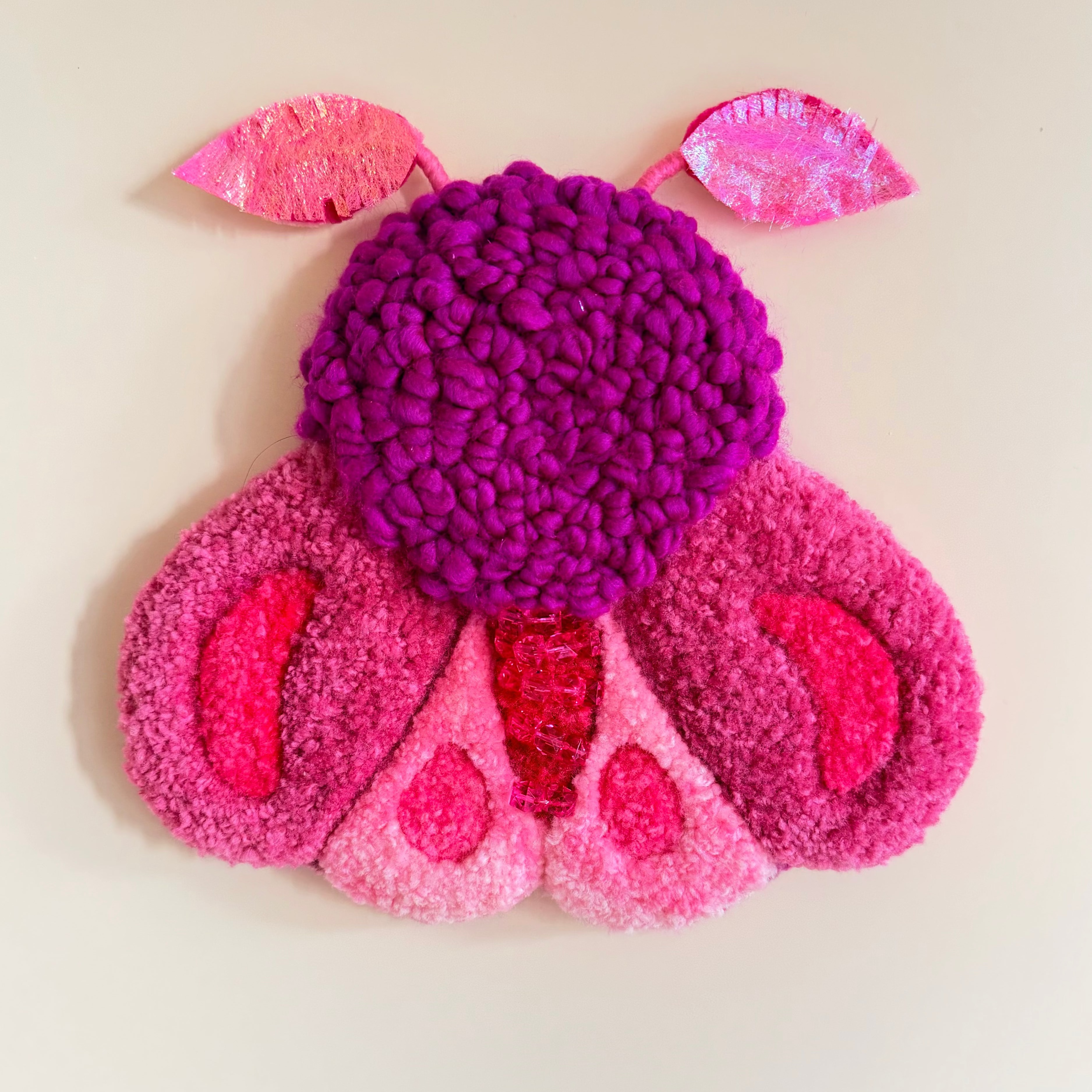 A colorful, textured wall hanging shaped like a stylized moth, made from punch needle, style yarn in shades of magenta, pink, and purple, mounted on a plain beige wall.